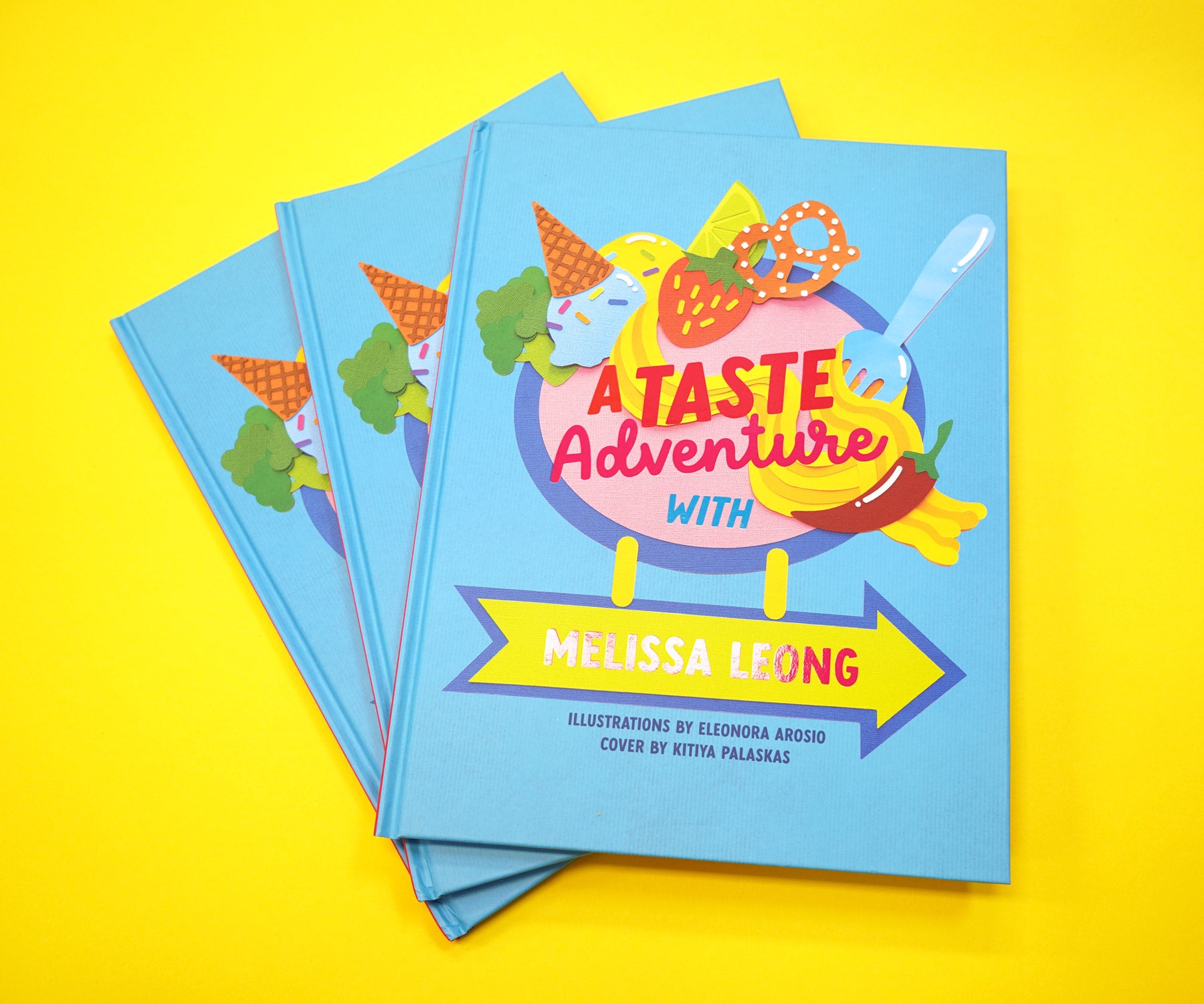 The book "A Taste Adventure" sits in a yellow background. The front cover features handmade craft based paper illustration of food by Kitiya Palaskas