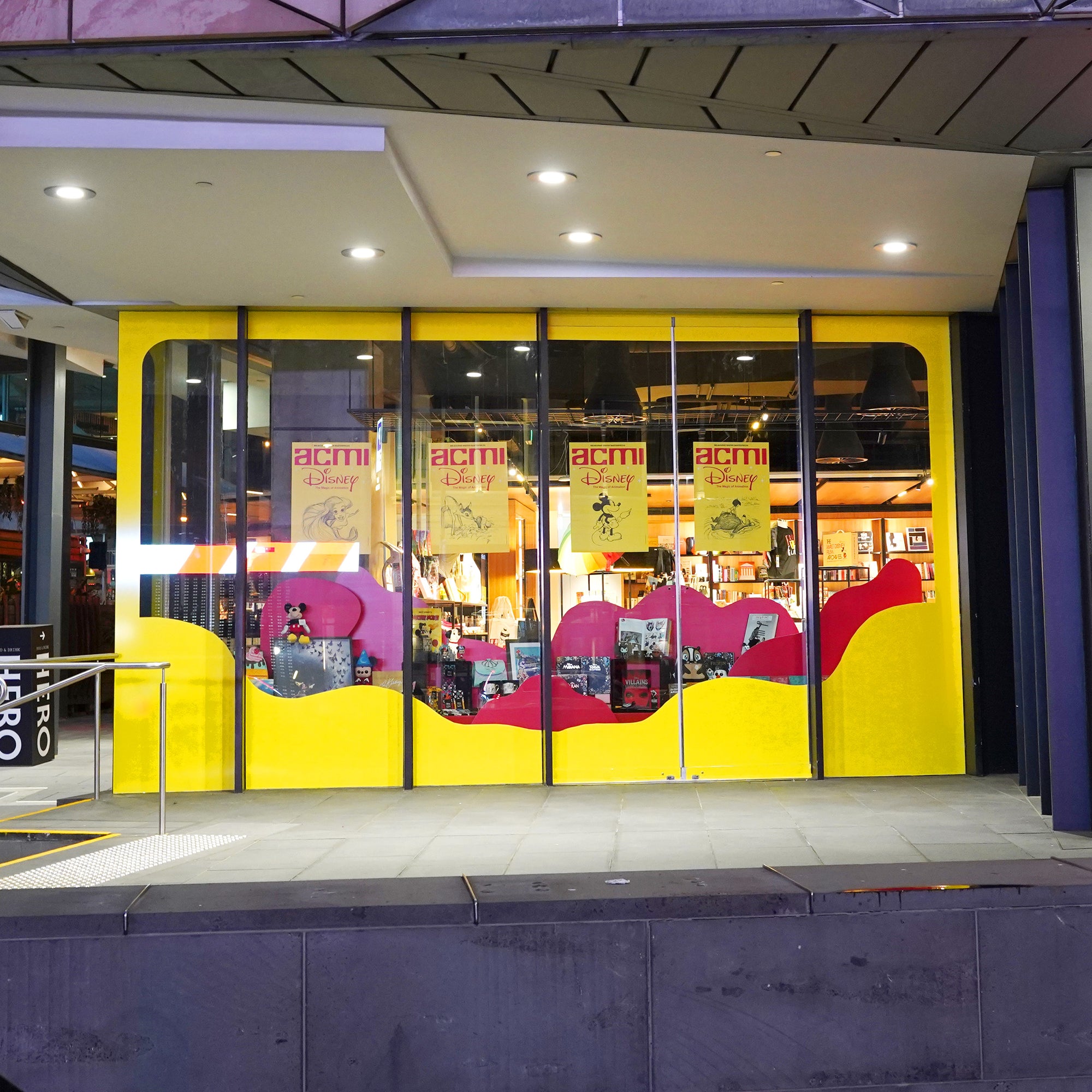 A Disney-themed retail window installation in the ACMI design shop, designed by Kitiya Palaskas Studio, with large red and white abstract window details, red abstract flat set pieces, and lots of Disney products. 