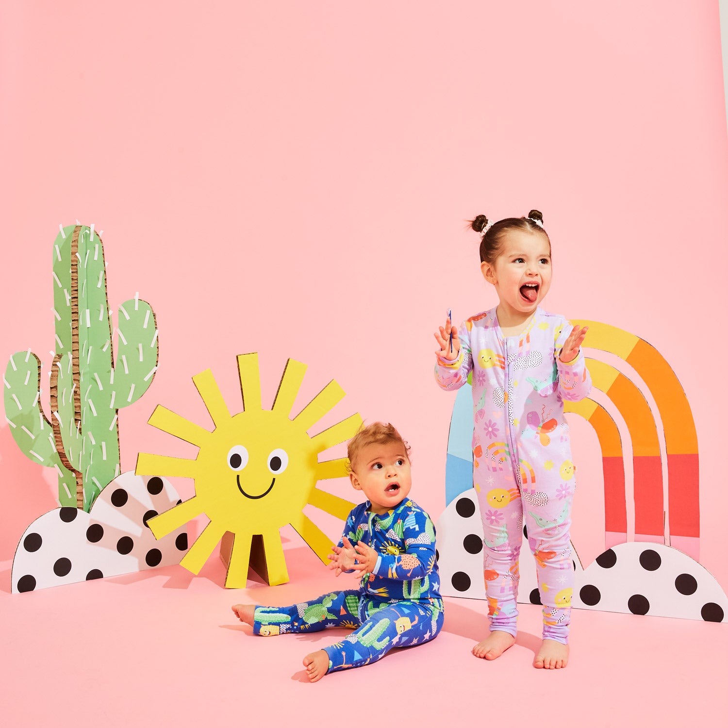 Children in patterned pyjamas play in a colourful background with custom Kitiya Palaskas cardboard props that include a sun, cactus, rainbow, clouds and butterfly