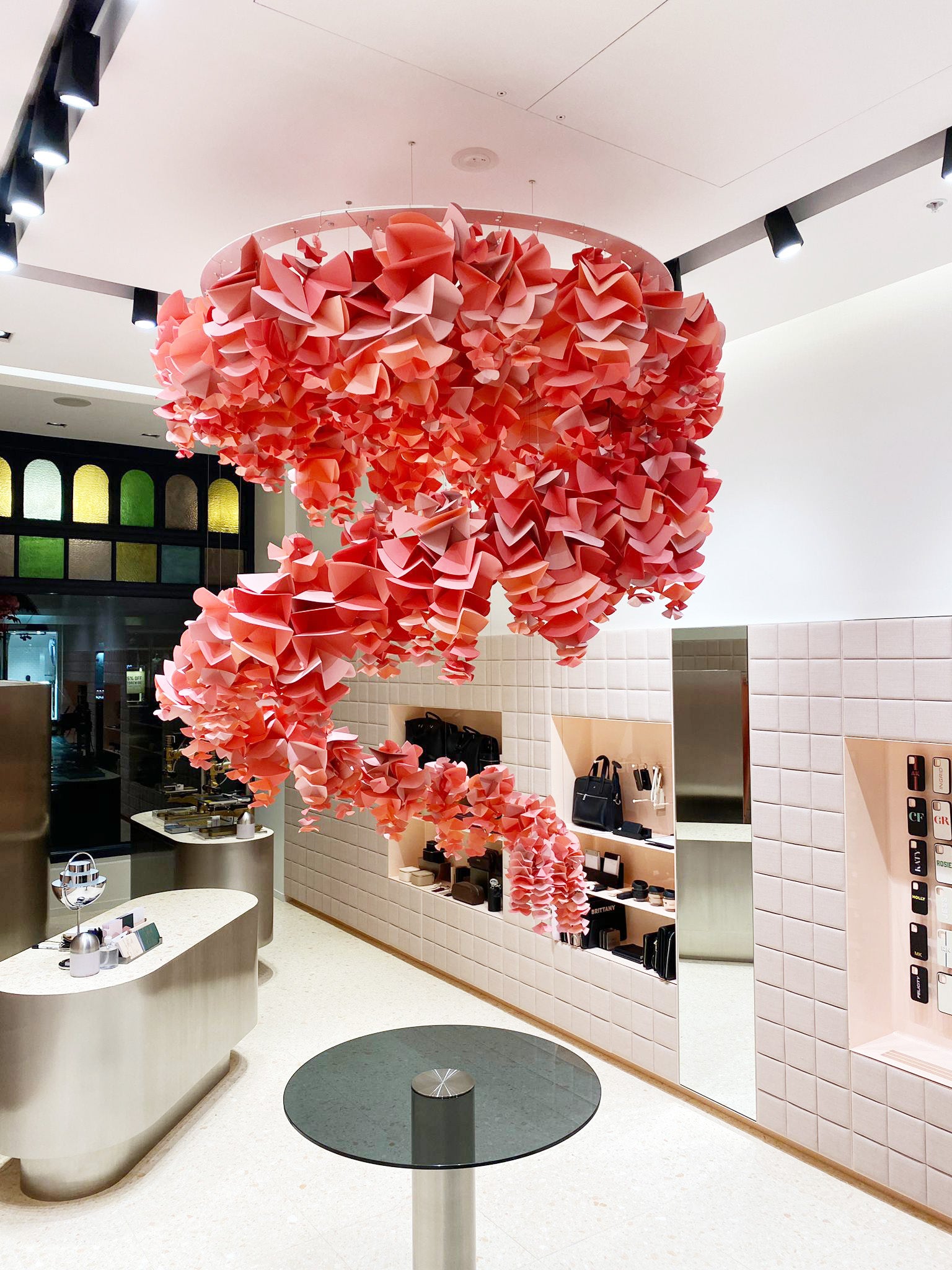 A large, pink, spiral-shaped papercraft petal installation hangs from the ceiling of a brightly lit retail store.