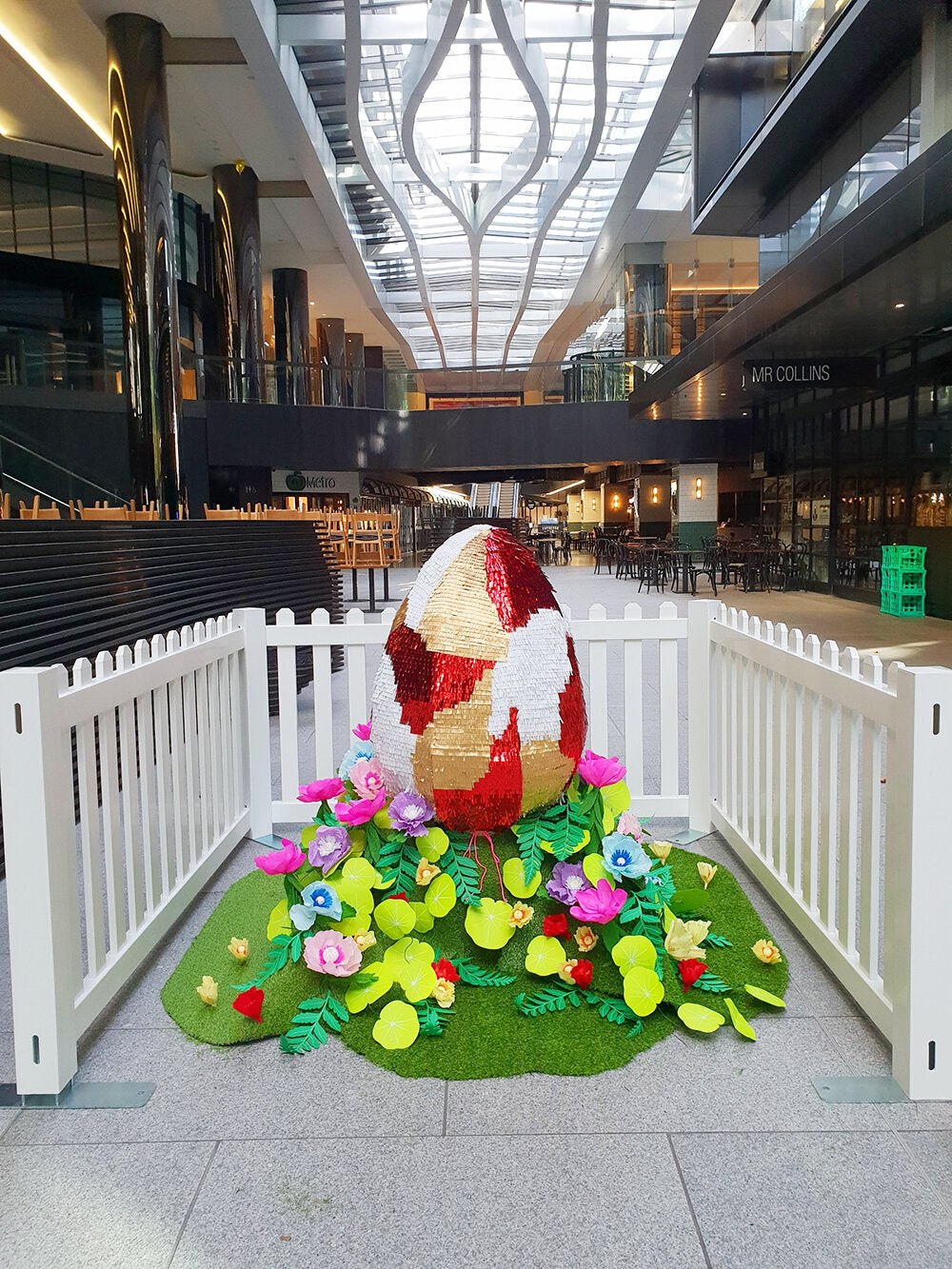 A giant red, gold and white piñata egg sits in a garden of colourful paper plants and flowers, surrounded by a white picket fence, inside a shopping centre.