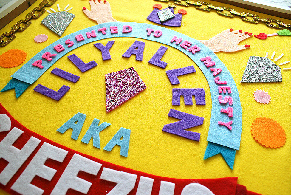 A colourful felt collaged artwork with a yellow background, and colourful motifs and words.