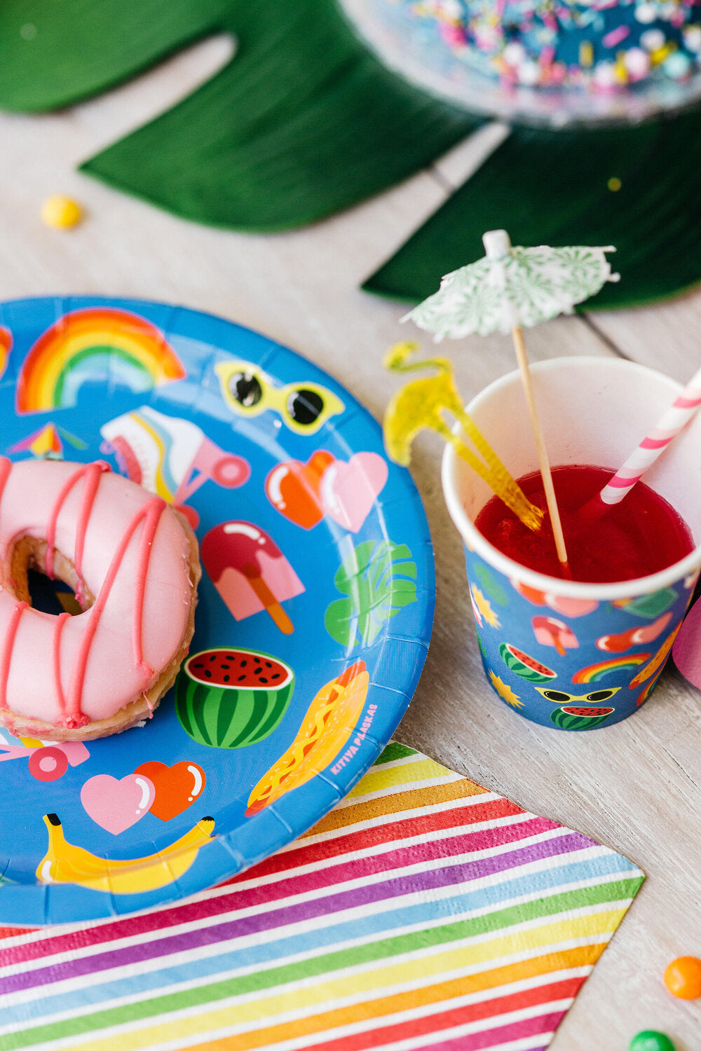 A collection of vibrant partyware in a party setting. The partyware includes plates, cups, party bags and piñatas and features a blue background, with tropical illustrated motifs. 