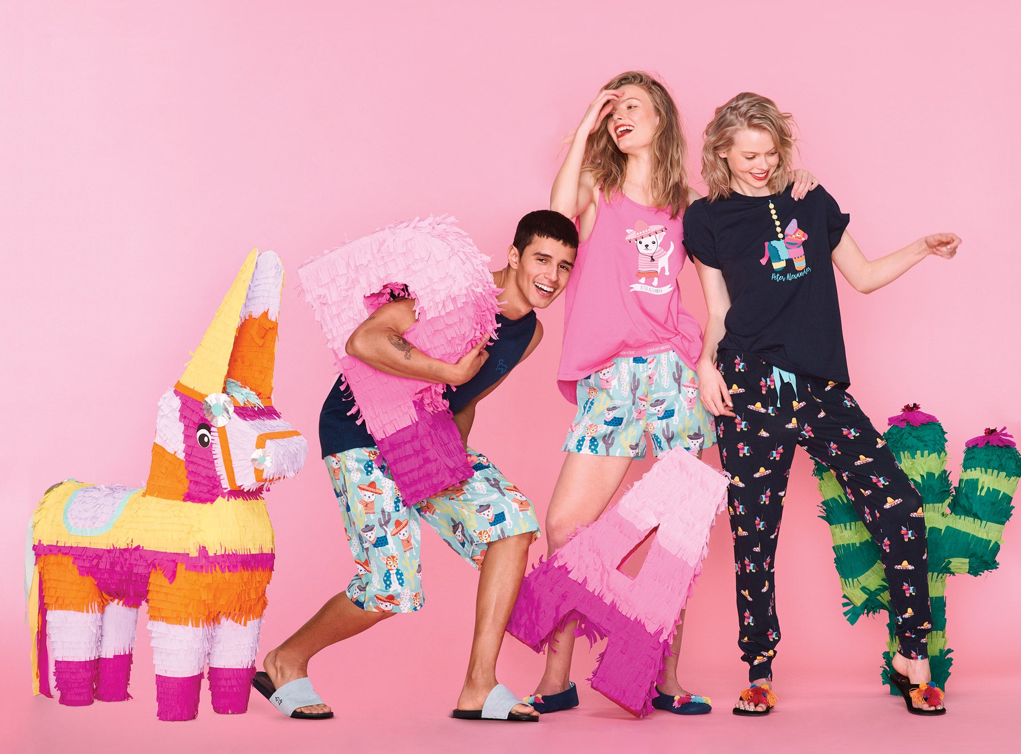 A group photo of 2 women and a man, posing against a pink background with a giant, colorful, oversized, handmade donkey piñata, two giant, oversized P and A shaped piñatas in shades of pink, and a giant green cactus piñata. 