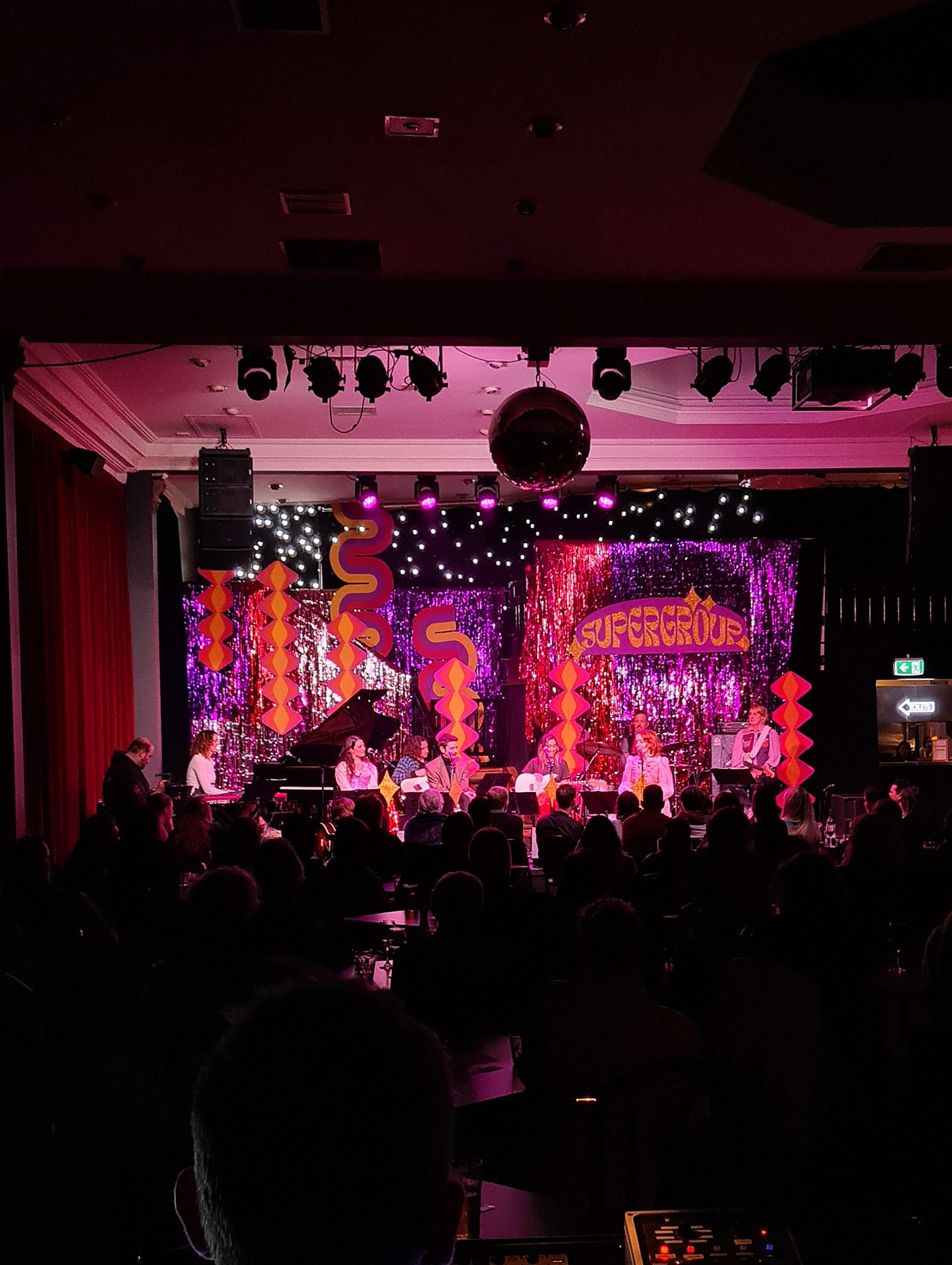 A live music venue featuring a colourful stage set design with shimmer curtains and retro-inspired cardboard cut-out set pieces. 