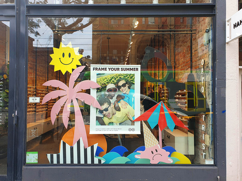 A colourful installation of cut-out shapes decorates. Bailey Nelson retail store window. The  window props include a giant pair of eyeglasses, stripy beach umbrella, sun with smiley face, black and white striped starburst shape, melted ice cream with sad 